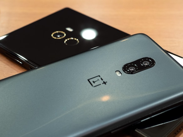 Two smartphones placed on a wooden surface, one is grey metallic with dual rear cameras and a brand logo, while the second is glossy black with a single camera and circular design around it.
