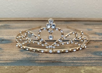 An ornate gold tiara adorned with sparkling clear gemstones, arranged in a symmetrical design with intricate loops and a prominent heart shape at its center. It rests on a wooden surface with a neutral background.