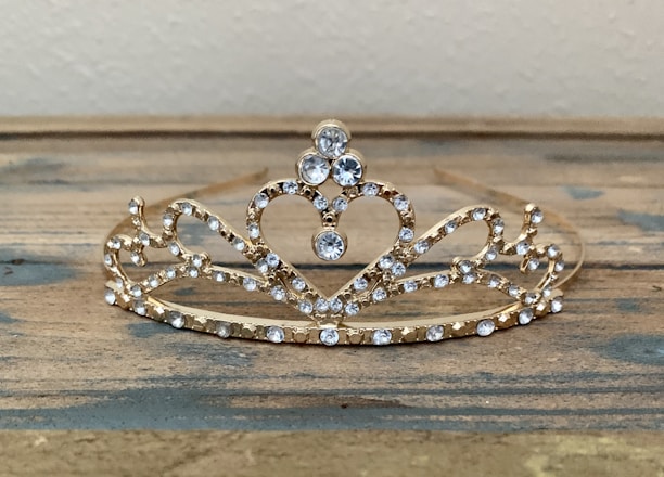 An ornate gold tiara adorned with sparkling clear gemstones, arranged in a symmetrical design with intricate loops and a prominent heart shape at its center. It rests on a wooden surface with a neutral background.