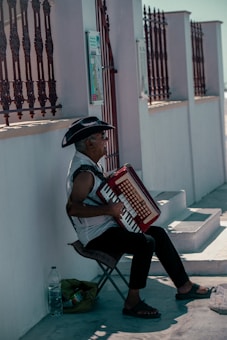 An elderly man wearing a cowboy hat sits on a folding chair outdoors, playing an accordion. He is next to a light-colored wall with decorative ironwork and steps leading upward. A large plastic bottle of water and a bag are placed beside him on the ground.