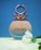 A perfume bottle with a unique metallic design at the top, against a blue background. The bottle is labeled 'Colors' and is positioned on a yellow surface. A white flower is placed next to it.