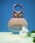 A perfume bottle with a unique metallic design at the top, against a blue background. The bottle is labeled 'Colors' and is positioned on a yellow surface. A white flower is placed next to it.
