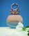 A perfume bottle with a unique metallic design at the top, against a blue background. The bottle is labeled 'Colors' and is positioned on a yellow surface. A white flower is placed next to it.