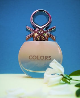 A perfume bottle with a unique metallic design at the top, against a blue background. The bottle is labeled 'Colors' and is positioned on a yellow surface. A white flower is placed next to it.