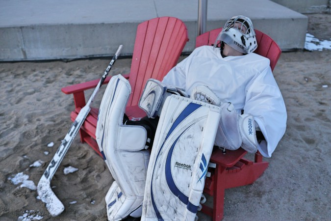 A hockey goalie in white pads takes a break in a red chair, a lighthearted look at players waiting to learn where they will play this season