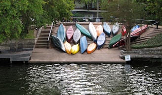 A collection of colorful kayaks is neatly arranged on a concrete ramp leading down to the water. The ramp is flanked by stone walls and railings, and surrounded by lush green trees. Seating is visible to the left near a small dock area. A sign reads 'Absolutely NO MO' near the water edge.