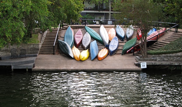A collection of colorful kayaks is neatly arranged on a concrete ramp leading down to the water. The ramp is flanked by stone walls and railings, and surrounded by lush green trees. Seating is visible to the left near a small dock area. A sign reads 'Absolutely NO MO' near the water edge.