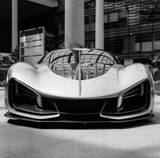 A sleek and futuristic sports car is positioned indoors, exhibiting a smooth body with aerodynamic curves and aggressive styling. The reflected light patterns enhance the metallic sheen on the car's surface. Surrounding elements include architectural features like large glass and steel structures, contributing to a modern atmosphere.