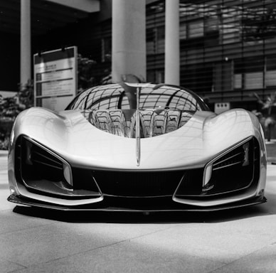 A sleek and futuristic sports car is positioned indoors, exhibiting a smooth body with aerodynamic curves and aggressive styling. The reflected light patterns enhance the metallic sheen on the car's surface. Surrounding elements include architectural features like large glass and steel structures, contributing to a modern atmosphere.