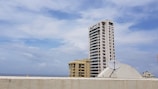 A tall residential building with multiple windows stands prominently against a partly cloudy sky. Below, a satellite dish is mounted on a rooftop, and the sea is visible in the distance. Other buildings are also present, contributing to an urban skyline.