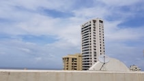 A tall residential building with multiple windows stands prominently against a partly cloudy sky. Below, a satellite dish is mounted on a rooftop, and the sea is visible in the distance. Other buildings are also present, contributing to an urban skyline.