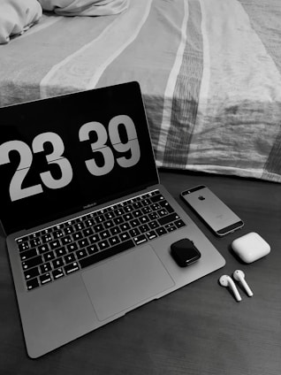 A workspace setup with a laptop displaying a digital clock showing 23:39. Next to the laptop, there is a smartphone, wireless earbuds with their charging case, and a small electronic device. The scene is set on a wooden table beside a neatly made bed with a patterned duvet.