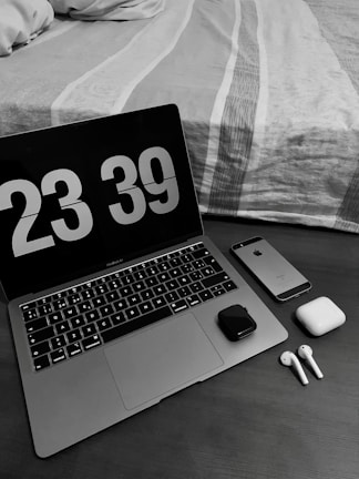A workspace setup with a laptop displaying a digital clock showing 23:39. Next to the laptop, there is a smartphone, wireless earbuds with their charging case, and a small electronic device. The scene is set on a wooden table beside a neatly made bed with a patterned duvet.