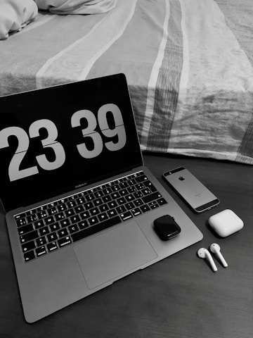 A workspace setup with a laptop displaying a digital clock showing 23:39. Next to the laptop, there is a smartphone, wireless earbuds with their charging case, and a small electronic device. The scene is set on a wooden table beside a neatly made bed with a patterned duvet.
