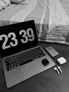 A workspace setup with a laptop displaying a digital clock showing 23:39. Next to the laptop, there is a smartphone, wireless earbuds with their charging case, and a small electronic device. The scene is set on a wooden table beside a neatly made bed with a patterned duvet.