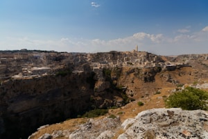 A historic town with stone buildings and a prominent tower sits atop a rocky hill. The area is surrounded by a rugged landscape with cliffs and sparse vegetation under a clear blue sky.