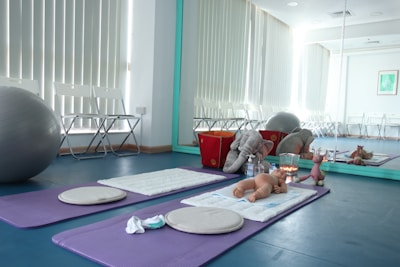 A room with yoga mats and soft pillows on the floor, along with a baby doll and stuffed animals, including an elephant and a giraffe. A large exercise ball and some folded chairs are situated against the wall, which features a tall mirror reflecting the interior. The setting appears serene and organized.