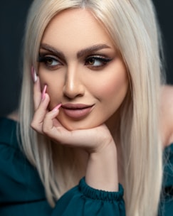 A woman with long blonde hair and subtle makeup rests her chin on her hand. She has striking, well-defined eyebrows and full lips. The nail polish on her manicured nails is a light pink color, and she is wearing a dark green off-shoulder top.