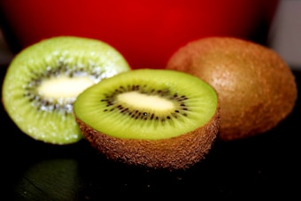 Two ripe kiwi fruits are prominently displayed. One is cut in half, showcasing the vibrant green flesh with tiny black seeds arranged around a white center. The other kiwi is whole, revealing the brown, fuzzy skin characteristic of this fruit. The background features a deep red color, adding contrast to the image.
