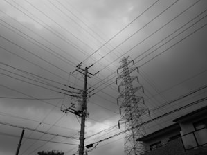 A complex network of power lines stretches across a cloudy sky. A utility pole and transmission tower dominate the scene, intersecting at various angles. The ground is partly visible with a building and some foliage.