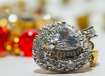 A sparkling heart-shaped diamond surrounded by smaller round diamonds on a silver setting, with an out-of-focus background featuring vibrant red and yellow hues from blurred ornaments.