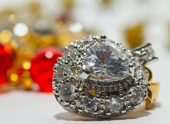 A sparkling heart-shaped diamond surrounded by smaller round diamonds on a silver setting, with an out-of-focus background featuring vibrant red and yellow hues from blurred ornaments.