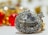 A sparkling heart-shaped diamond surrounded by smaller round diamonds on a silver setting, with an out-of-focus background featuring vibrant red and yellow hues from blurred ornaments.
