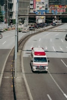 An ambulance is driving on a city road, specifically in the rightmost lane. Surrounding the vehicle are multiple lanes, some carrying regular traffic. In the background, there are buildings adorned with various colorful advertisements and signs, likely indicating a bustling urban area.