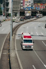 An ambulance is driving on a city road, specifically in the rightmost lane. Surrounding the vehicle are multiple lanes, some carrying regular traffic. In the background, there are buildings adorned with various colorful advertisements and signs, likely indicating a bustling urban area.