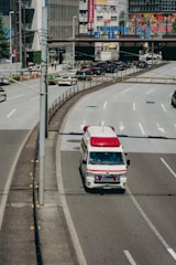 An ambulance is driving on a city road, specifically in the rightmost lane. Surrounding the vehicle are multiple lanes, some carrying regular traffic. In the background, there are buildings adorned with various colorful advertisements and signs, likely indicating a bustling urban area.