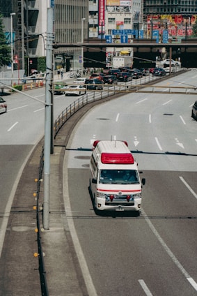 An ambulance is driving on a city road, specifically in the rightmost lane. Surrounding the vehicle are multiple lanes, some carrying regular traffic. In the background, there are buildings adorned with various colorful advertisements and signs, likely indicating a bustling urban area.