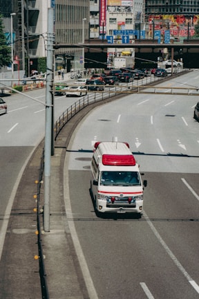 An ambulance is driving on a city road, specifically in the rightmost lane. Surrounding the vehicle are multiple lanes, some carrying regular traffic. In the background, there are buildings adorned with various colorful advertisements and signs, likely indicating a bustling urban area.