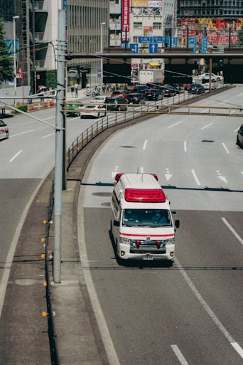 An ambulance is driving on a city road, specifically in the rightmost lane. Surrounding the vehicle are multiple lanes, some carrying regular traffic. In the background, there are buildings adorned with various colorful advertisements and signs, likely indicating a bustling urban area.