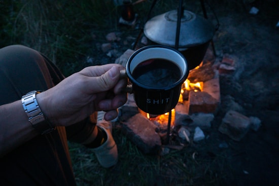 A person holds a mug with dark liquid in front of a campfire. The mug has a humorous exclamation written on it. There is a cooking pot on a stand over the fire, surrounded by bricks, with grass visible nearby.
