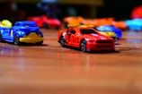 Toy cars are scattered on a wooden surface, with a red sports car and blue service truck prominently in focus. The background features blurred, colorful toy cars creating a playful and dynamic atmosphere.