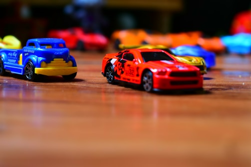 Toy cars are scattered on a wooden surface, with a red sports car and blue service truck prominently in focus. The background features blurred, colorful toy cars creating a playful and dynamic atmosphere.