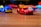 Toy cars are scattered on a wooden surface, with a red sports car and blue service truck prominently in focus. The background features blurred, colorful toy cars creating a playful and dynamic atmosphere.