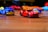 Toy cars are scattered on a wooden surface, with a red sports car and blue service truck prominently in focus. The background features blurred, colorful toy cars creating a playful and dynamic atmosphere.