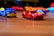 Toy cars are scattered on a wooden surface, with a red sports car and blue service truck prominently in focus. The background features blurred, colorful toy cars creating a playful and dynamic atmosphere.