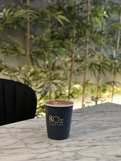 A cup of coffee with latte art sits on a marble table. The background features tall green plants, creating a natural and relaxing setting.