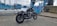 A motorcycle is parked on a paved area near a closed blue metal gate with a sign in Welsh. In the background, there is an industrial structure with steel beams and towers, possibly a bridge. The sky is overcast.