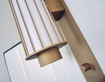 A metallic wall-mounted light fixture with a cylindrical design is prominently displayed. The fixture features vertical bars and a white diffuser, giving it a stylish and modern look. The background includes a textured white surface and a framed section, adding to the contemporary aesthetic.