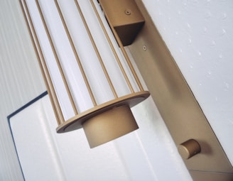 A metallic wall-mounted light fixture with a cylindrical design is prominently displayed. The fixture features vertical bars and a white diffuser, giving it a stylish and modern look. The background includes a textured white surface and a framed section, adding to the contemporary aesthetic.