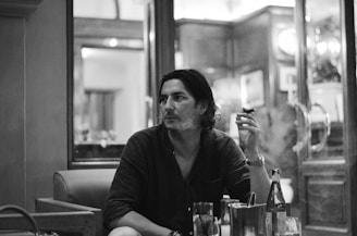 A person is sitting in a dimly lit indoor setting, holding a cigar. The background features wooden panels and mirrors, suggesting a lounge or a sophisticated bar environment. The atmosphere appears relaxed and introspective.