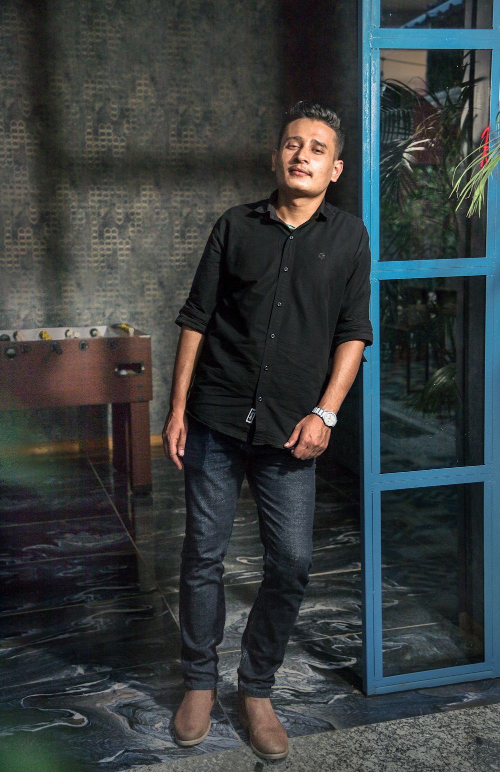 Foto hombre con camisa negra y pantalones negros parado junto a una puerta  de vidrio con marco de madera azul – Imagen Nepal gratis en Unsplash