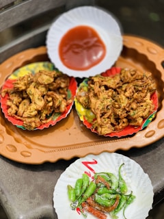 Two colorful paper plates filled with crispy fried snacks are placed on an orange tray. Additionally, there is a smaller white paper plate containing fried green chili peppers and salt. A separate white paper dish holds red dipping sauce.