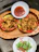Two colorful paper plates filled with crispy fried snacks are placed on an orange tray. Additionally, there is a smaller white paper plate containing fried green chili peppers and salt. A separate white paper dish holds red dipping sauce.