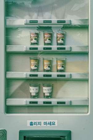 A vending machine displays various cups of instant coffee and tea. The cups are arranged in three rows, with different labels and prices marked on them in Korean. The price labels indicate values such as 400 won and 300 won. The cups in the top two rows show different flavors of Maxim coffee, while the bottom row has cups labeled with handwritten tags. The machine casing and backdrop are in muted colors with shadows visible on the surface.