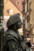 A detailed figure of a street performer, covered entirely in dark gray paint to resemble a statue, wearing historical attire with a hat and standing against an urban backdrop with blurred buildings.