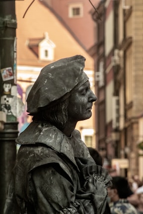 A detailed figure of a street performer, covered entirely in dark gray paint to resemble a statue, wearing historical attire with a hat and standing against an urban backdrop with blurred buildings.